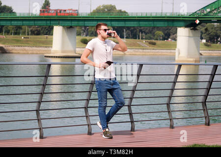 Man leaning on the fence using tablet and headphones, enjoying his surroundings by the river and old steel, green colored bridge. Urban, city. Stock Photo