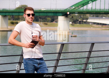 Man leaning on the fence using tablet and headphones, enjoying his surroundings by the river and old steel, green colored bridge. Urban, city. Stock Photo