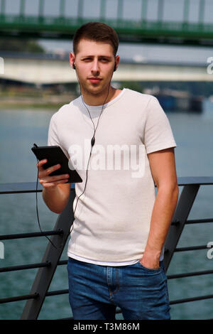 Man leaning on the fence using tablet and headphones, enjoying his surroundings by the river and old steel, green colored bridge. Urban, city. Stock Photo