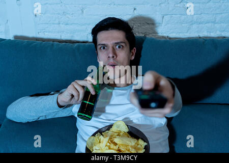 Lifestyle portrait of young man feeling scared and shocked making fear, anxiety gestures while watching TV, holding remote control and drinking a beer Stock Photo