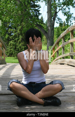 Beautiful dark haired girl sitting on a wooden bridge and wearing a white T shirt and blue jean cut offs and hiding her face Stock Photo