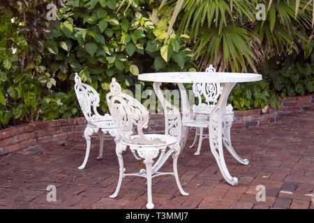 ornate white wrought iron table and chair set on brick patio in tropical garden Stock Photo