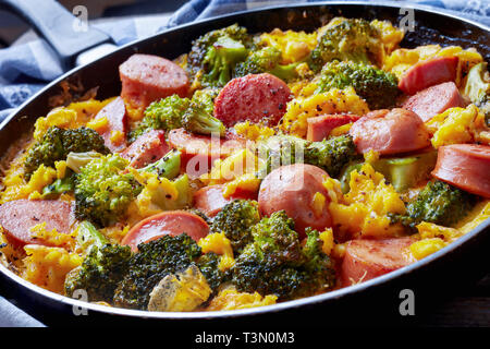 delicious hot scrambled eggs with broccoli and sausages in a frying pan on an old wooden table with kitchen towel, horizontal view from above, close-u