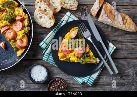 omelette with broccoli and sausages in a skillet and served on a plate on an old grey rustic wooden table with sliced crusty whole grain French baguet