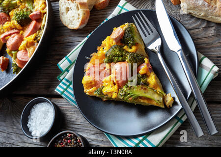 frittata with broccoli and sausages served on a black plate on an old grey rustic wooden table with sliced crusty whole grain French baguette, view fr Stock Photo