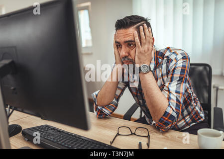 Handsome serious pensive businessman is working on computer in the office. He is worried about something showing on the computer. Stock Photo