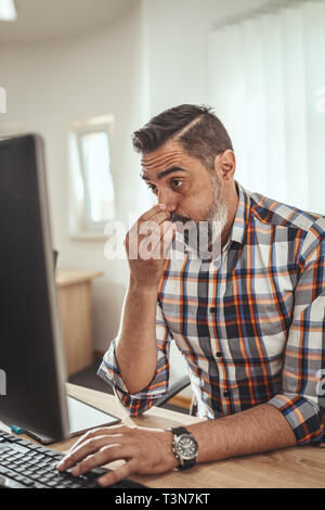 Young handsome serious pensive businessman is working on computer in the office. He is worried about something showing on the computer. Stock Photo