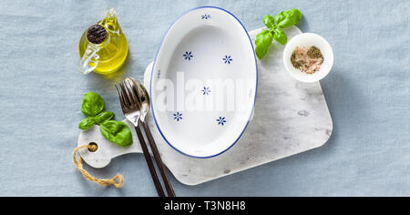 banner of empty ceramic white baking dish on a marble cutting board and cutlery on a blue linen tablecloth with olive oil, spices and basil leaves. ho Stock Photo