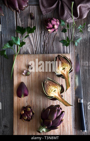 Flat lay with fresh red artishokes, whole and halved, on cutting board on rustic wooden background Stock Photo