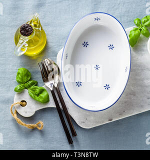 empty ceramic white baking dish on a marble cutting board and cutlery on a blue linen tablecloth with olive oil, spices and basil leaves. home cooking Stock Photo