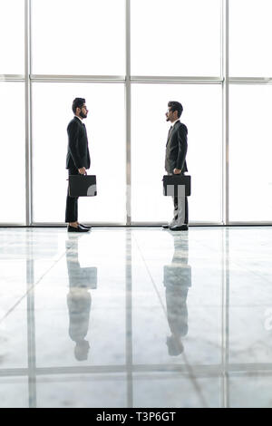 Two businessmen deep in discussion together while standing in an office boardroom with windows overlooking the ocean and city Stock Photo