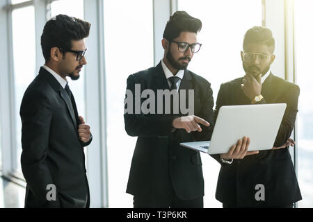 silhouettes of three asian corporate executives standing in front of windows discussing business using laptop computer. Stock Photo