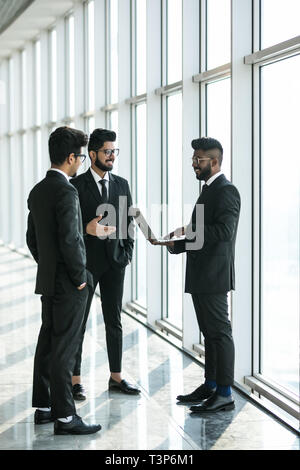 silhouettes of three asian corporate executives standing in front of windows discussing business using laptop computer. Stock Photo