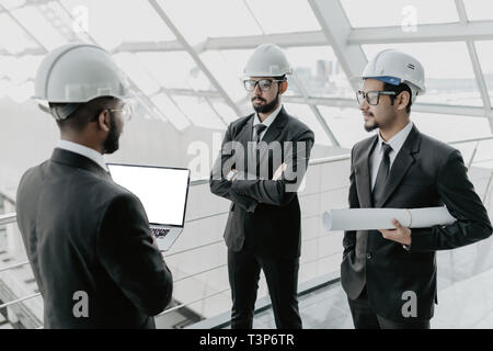 Closeup of three smiling diverse business people wearing helmets and standing outdoors with building in background. Architects concept. Stock Photo