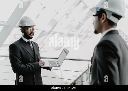 Closeup of three smiling diverse business people wearing helmets and standing outdoors with building in background. Architects concept. Stock Photo