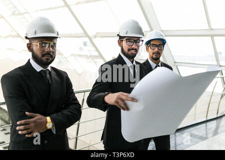 Qualified young architects are standing and discussing plan of building. They are looking with interest at blueprint while foreman is pointing finger  Stock Photo