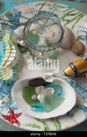 This is a beach theme still life of sea glass, a ceramic sea horse ornament, small glass jar with wired beads, some shells a fishing lure on a plate Stock Photo