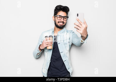 Smiling young guy taking selfie photo on smartphone. Indian man using digital device. Selfie photo concept. Isolated front view on white background. Stock Photo