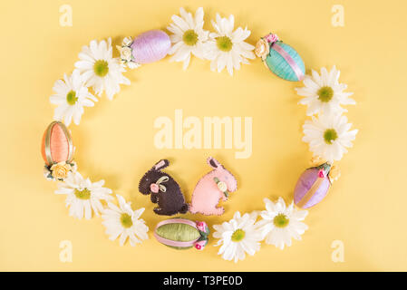 Wreath of daisies, Easter eggs, and Easter bunnies on solid pastel yellow background Stock Photo