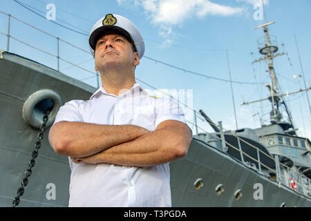 Captain standing in dock before warship and  looking ahead. A sailor officer in white uniform stands beside battleship. Stock Photo