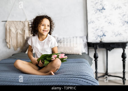 A little pretty curly girl sitting on her bed with tulips in her arms. Stock Photo