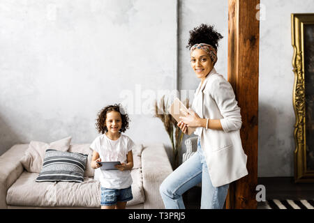 A cute mother and daughter talk and look at the tablet while standing in the living room Stock Photo