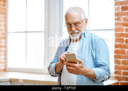 Cheerful senior grandfather using phone standing near the window at home Stock Photo