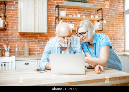 Beautiful senior couple in blue shirts sitting together with laptop on the kitchen at home Stock Photo