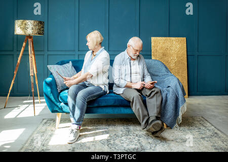 Offended senior man and woman sitting back to each other, using digital gadgets on the couch at home Stock Photo