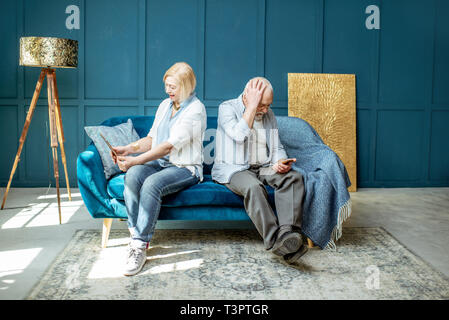 Offended senior man and woman sitting back to each other, using digital gadgets on the couch at home Stock Photo