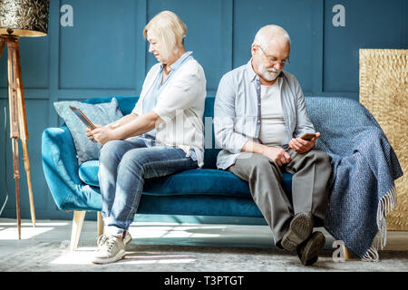 Offended senior man and woman sitting back to each other, using digital gadgets on the couch at home Stock Photo