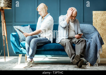 Offended senior man and woman sitting back to each other, using digital gadgets on the couch at home Stock Photo