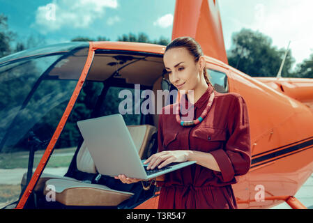 Pretty dark-haired busy woman standing with laptop and typing on it Stock Photo