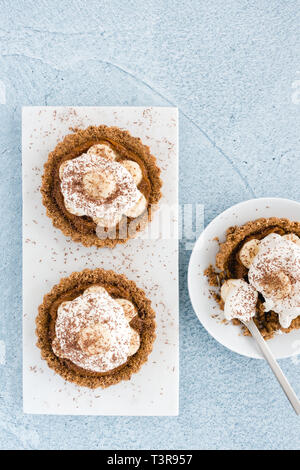 Top view of homemade Banoffee Pie Tartlets on white marble board and white porcelain plate with cake fork. Shot on blue cement background. Stock Photo