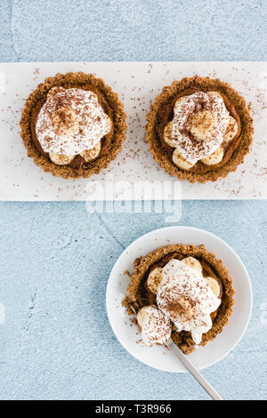 Homemade Banoffee Pie tartlets with salted caramel, banana slices, whipping cream decorated with chocolate on white marble chopping board and plate on Stock Photo