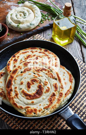 Chinese scallion Pancakes in a skillet with an uncooked pancake, sesame oil in a bottle, soy sauce and ingredients on a wooden table, vertical view fr Stock Photo
