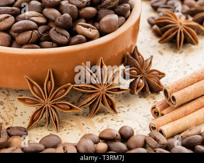 Black coffee beans lying on a clay dish. Cinnamon sticks and star anise star near the saucer of coffee beans on a cork board. Stock Photo