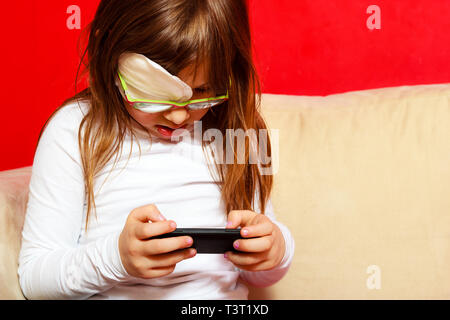 Technology generation. Little girl child in glasses with medicine plaster playing games on smartphone mobile phone at home Stock Photo