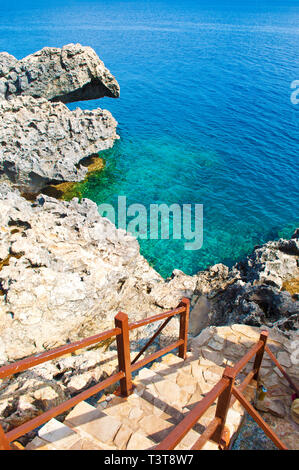Stone stairs with brown railings leading down to deep blue transparent azure water among rugged rocks near Cape Greco. Amazing seascape. Warm and sunn Stock Photo