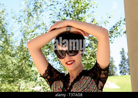 Portrait of a beautiful, dark haired lady wearing sunglasses in the park on a bright, sunny day. Hands over her head. Stock Photo