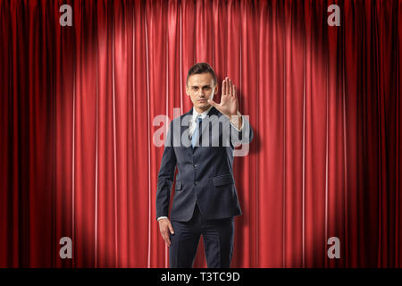 Front view of businessman standing in spotlight against red stage curtain looking at camera and holding out hand with open palm. Stock Photo