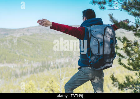 A male tourist hiker with a large backpack stands on a cliff stone in front of a green valley with his arms outstretched. The concept of freedom in tr Stock Photo