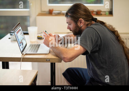 Young man with long hair using a phone while sitting in front of a working laptop at home. Social break is required for good communication Stock Photo