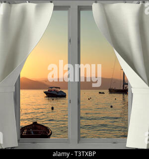Sunset with golden glow in the sky over boats at Portofino, Italy viewed through a window with white drapes pulled aside with reflection on the water Stock Photo