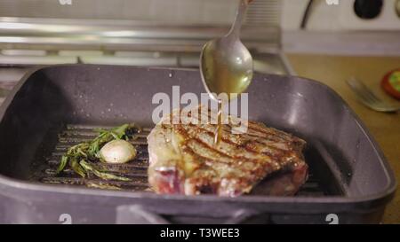 roasting of a beef steak with rosemary and garlic on a hot black pan and pouring oil on it by the spoon in unrecognizable man chef's hand, close up Stock Photo