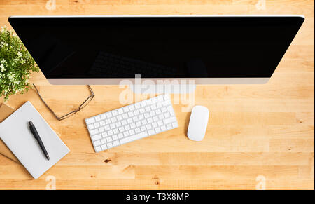 Modern desktop computer. Top view of wooden office desk with large desktop computer, keyboard, mouse, notebook, pen, glasses and plant. Modern office. Stock Photo