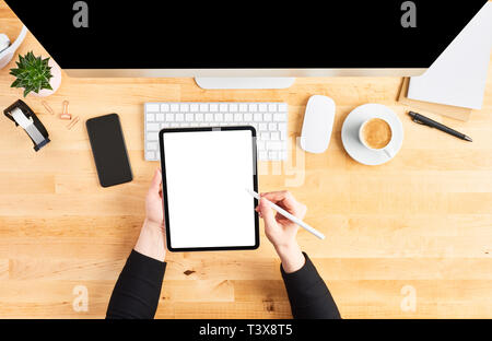 Female hands holding digital tablet and pencil. Top view of wooden office desk with tablet, pencil, large desktop computer, keyboard, mouse, smartphon Stock Photo