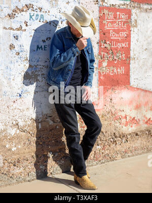 Old cowboy wearing cowboy boots and a hat leans up against a wall in a small southwestern town in Arizona. Stock Photo