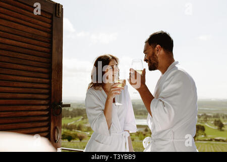 Smiling man and woman holding a glass of wine and talking to each other. Couple in bathrobe standing in balcony of house enjoying a glass of wine. Stock Photo