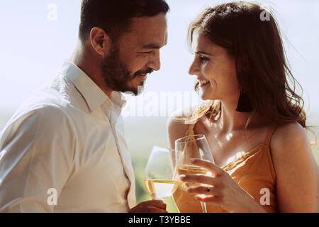 Happy man and woman standing together holding wine and looking at each other. Romantic couple toasting glasses of white wine on a date. Stock Photo